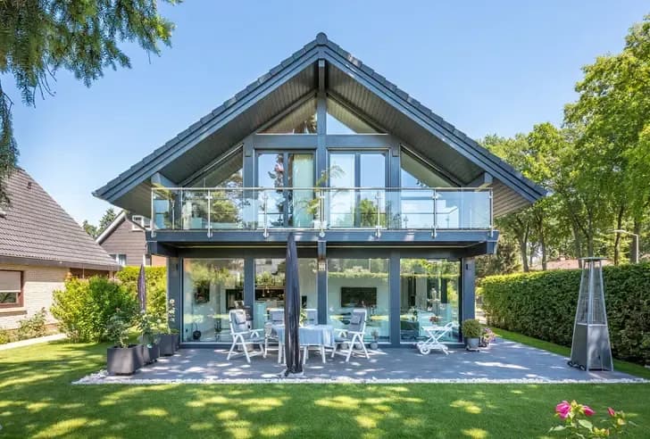 Modernes Haus mit großer Glasfront und Balkon Tisch mit weißen Gartenstühlen auf Terrasse und grüner Rasenfläche