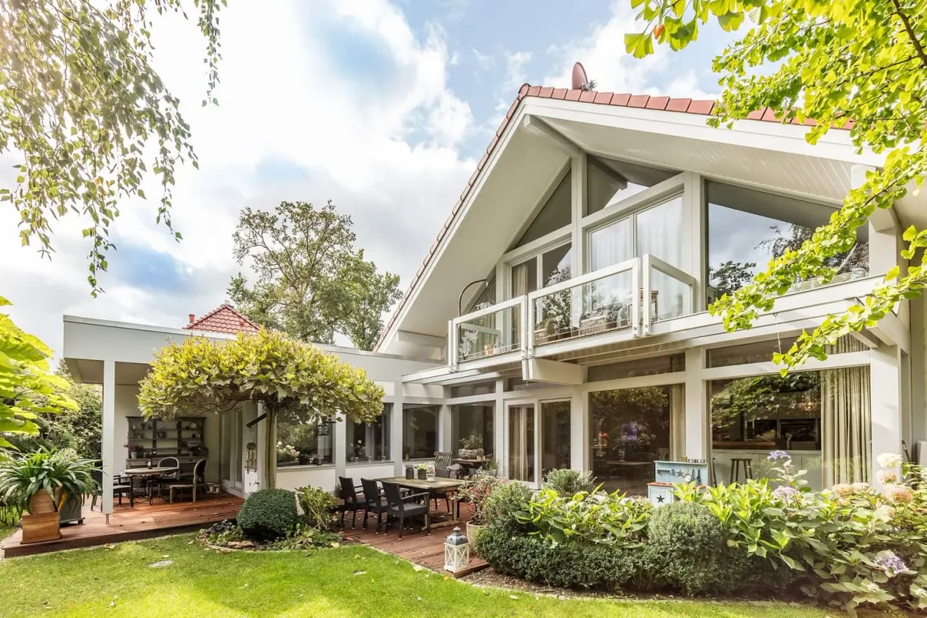 Modernes Einfamilienhaus mit weißer Holzverkleidung, Glasbalkon, Terrasse und üppigem grünen Garten bei Tageslicht