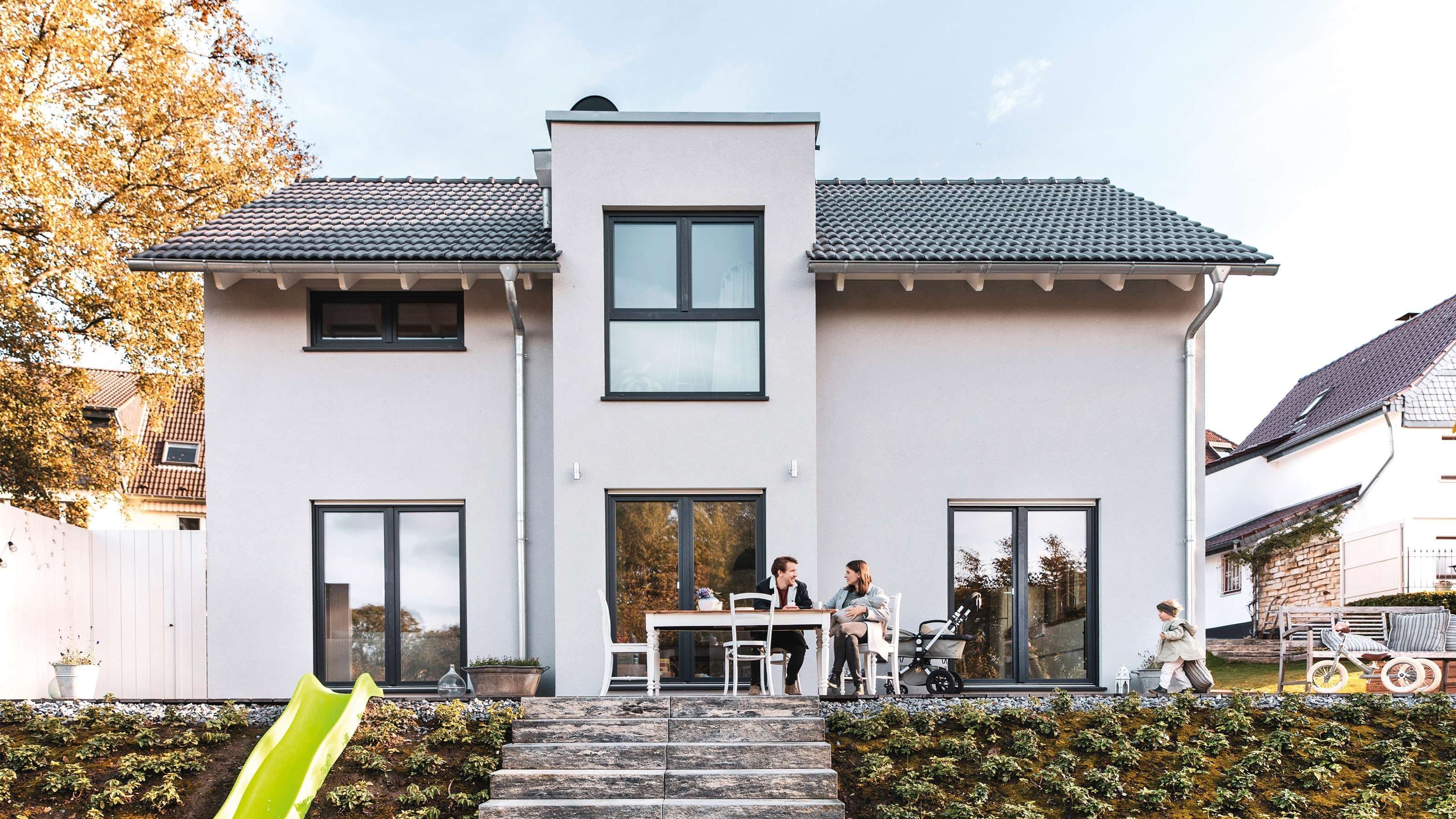 Modernes Einfamilienhaus mit grauem Dach, Terrasse, Garten und Familie beim Frühstück im Freien – stilvolles, energieeffizientes Hausdesign.